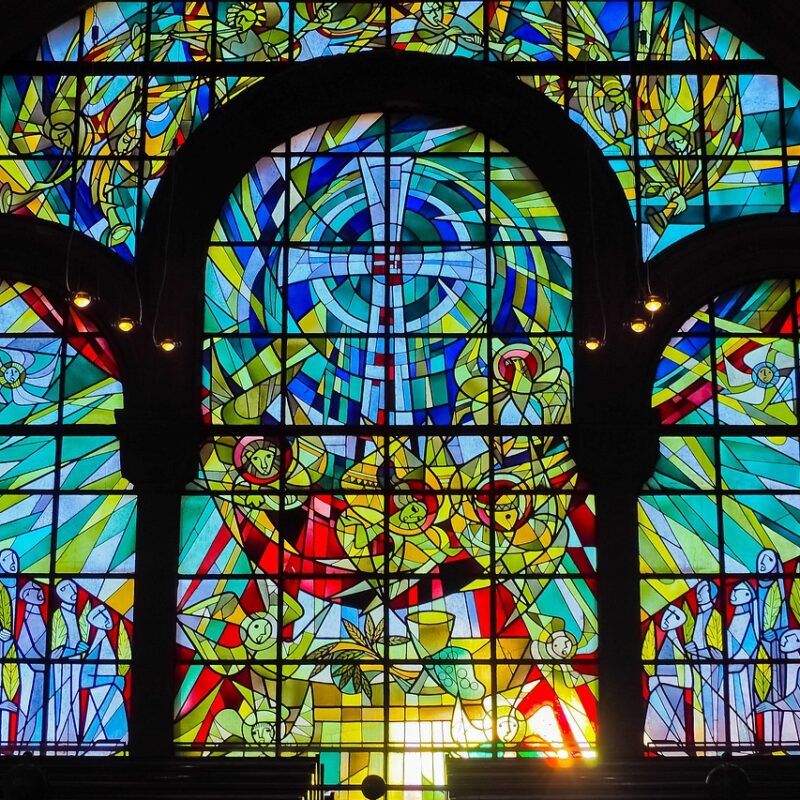 Colorful stained-glass window centered on a cross, with Eucharistic symbols and gathered figures.